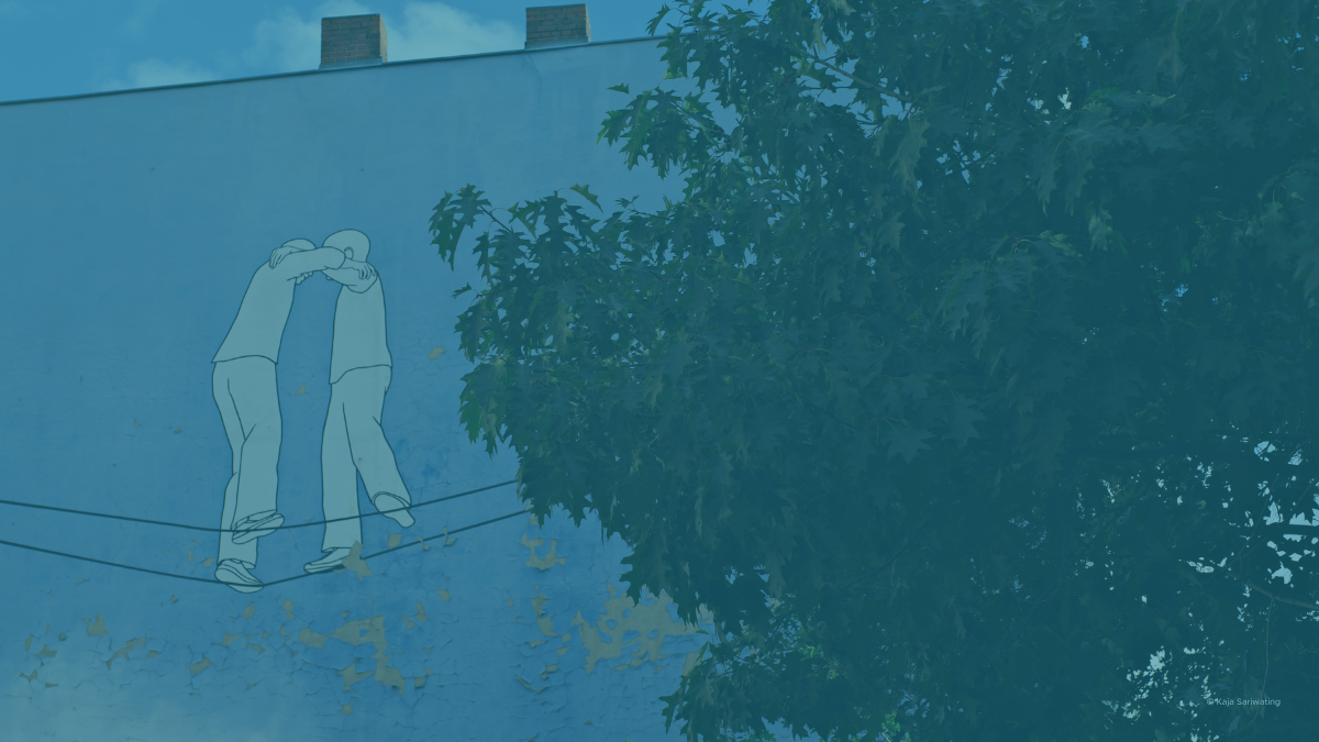 Streetart van Kaja Sariwating. Twee mensen die samen balanceren op 2 slappe koorden. Ze ondersteunen elkaar.